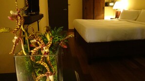 A hotel room with a bed, a wooden wardrobe, a desk with a lamp, and a glass vase with a plant.