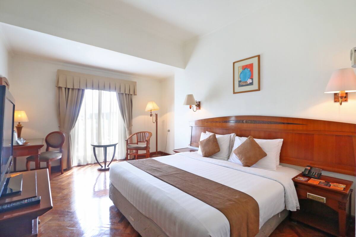 A hotel room with a large bed, bedside lamps, a desk with a computer, a chair, and a window with curtains.