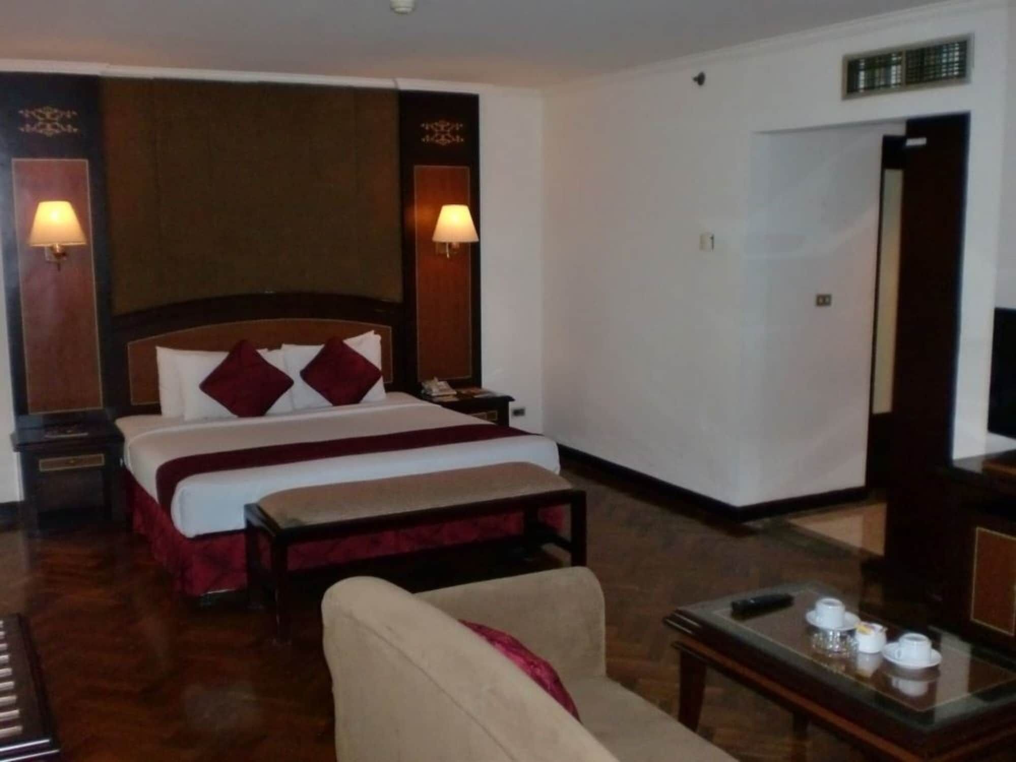 A hotel room with a large bed, a bedside table, a chair, a small table with cups, and a TV.