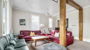 A living room with a grey sofa, a red leather sofa, a wooden coffee table, and a grey ottoman. There are two windows with white shutters, a lamp, and a guitar leaning against the wall. - Verandahs Parkside Lodge - Hostel (Auckland)