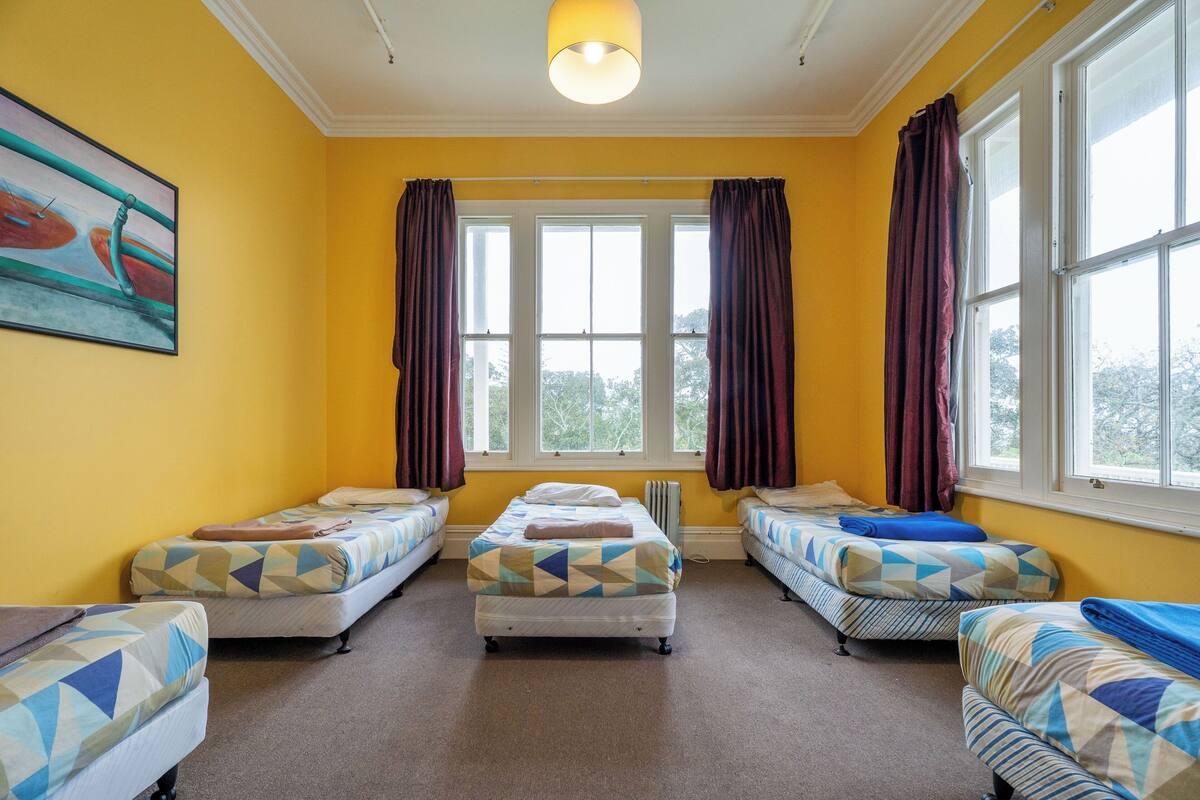 A room with four single beds, a radiator, and a large window with curtains.