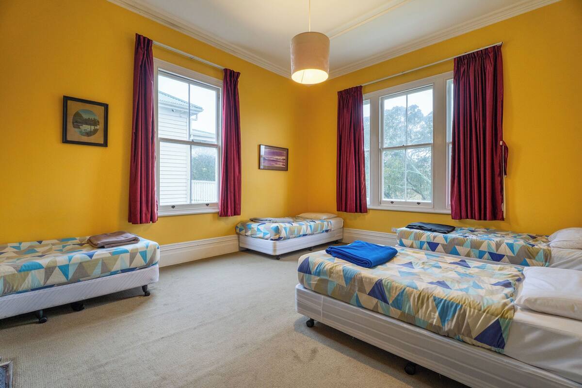 A room with three beds, yellow walls, and large windows with red curtains.
