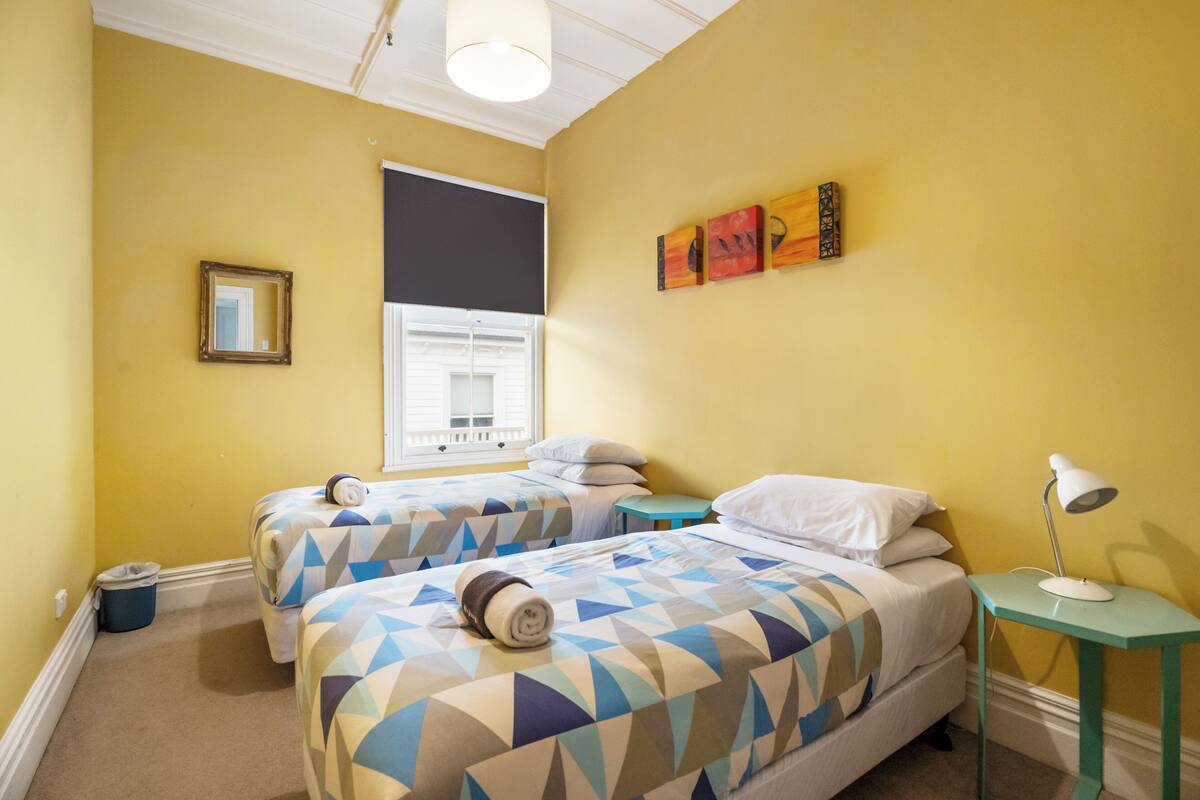 A room with two beds, a window, a small table with a lamp, and artwork on the wall.