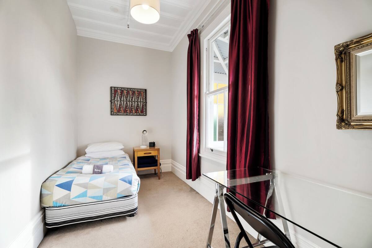 A bedroom with a bed, a bedside table, a lamp, a window with curtains, and a wall-mounted mirror.