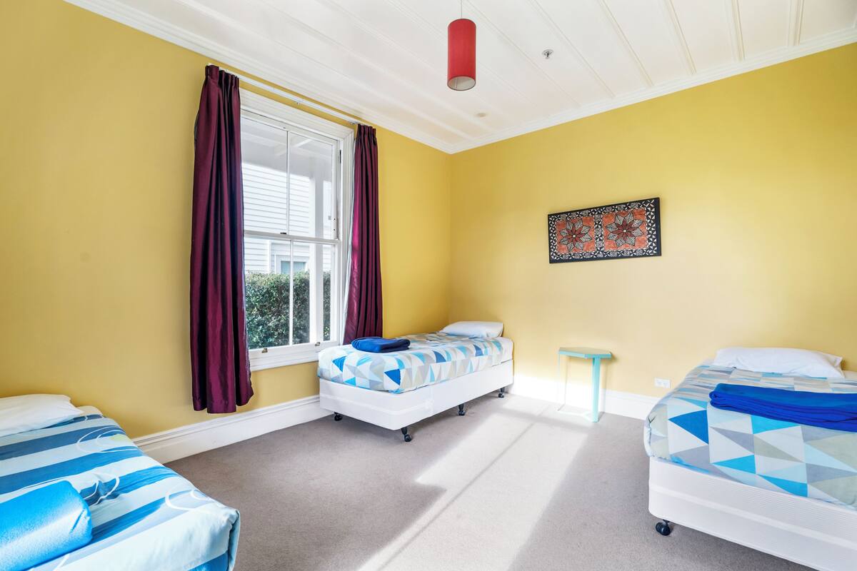 A room with two beds, a window with curtains, a small table, and a ceiling light.