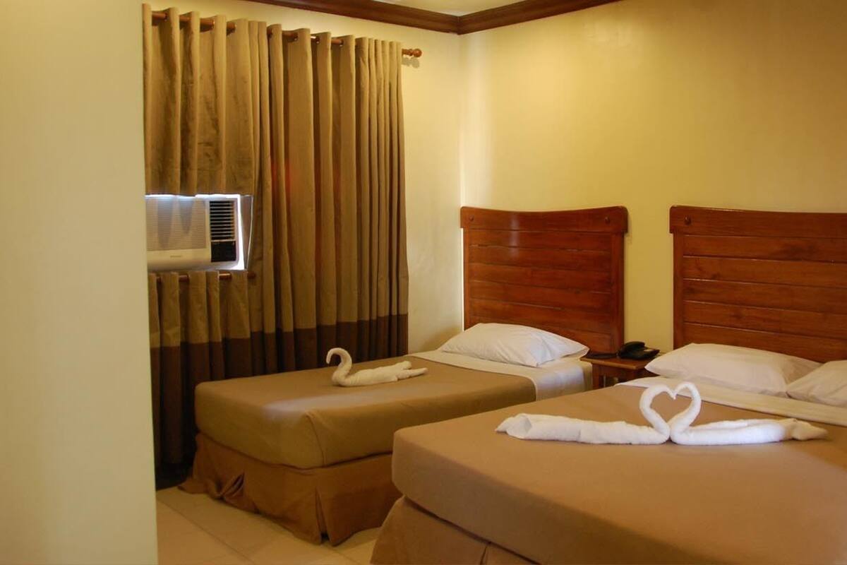 A hotel room with two single beds, wooden headboards, beige bedding, a curtain, and an air conditioning unit.