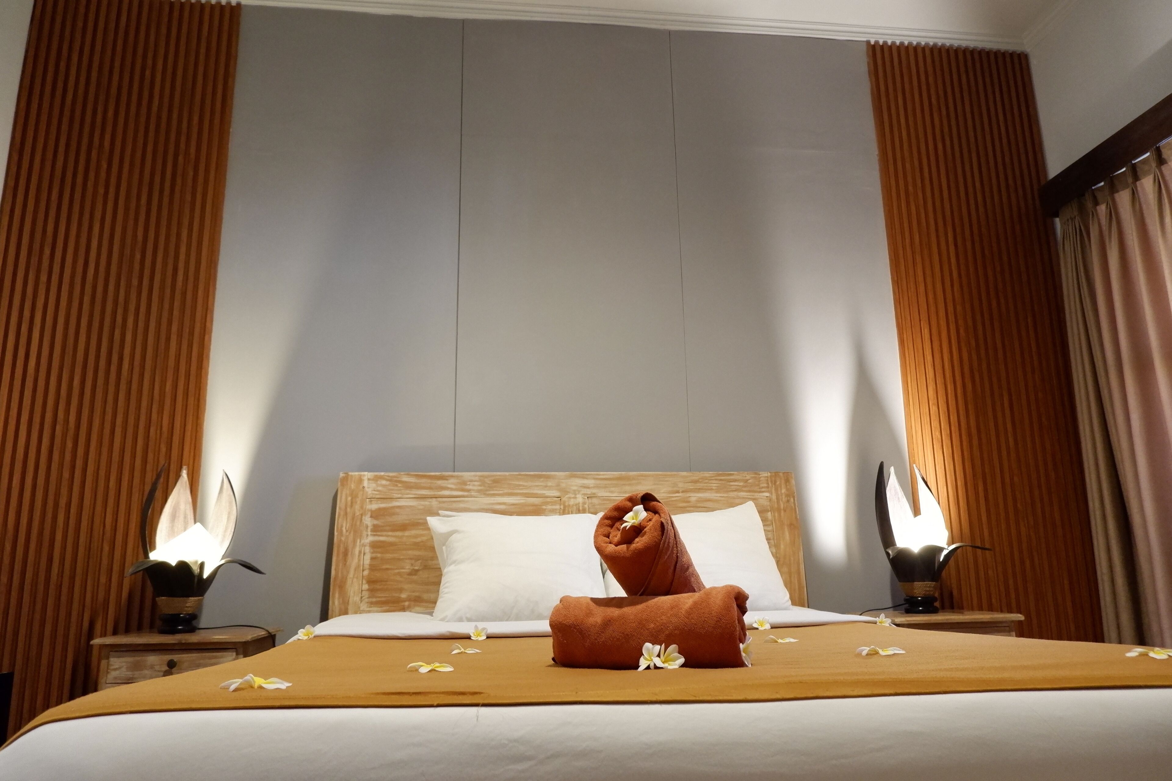 A neatly made bed with a brown towel and white flowers, wooden headboard, and two bedside lamps.
