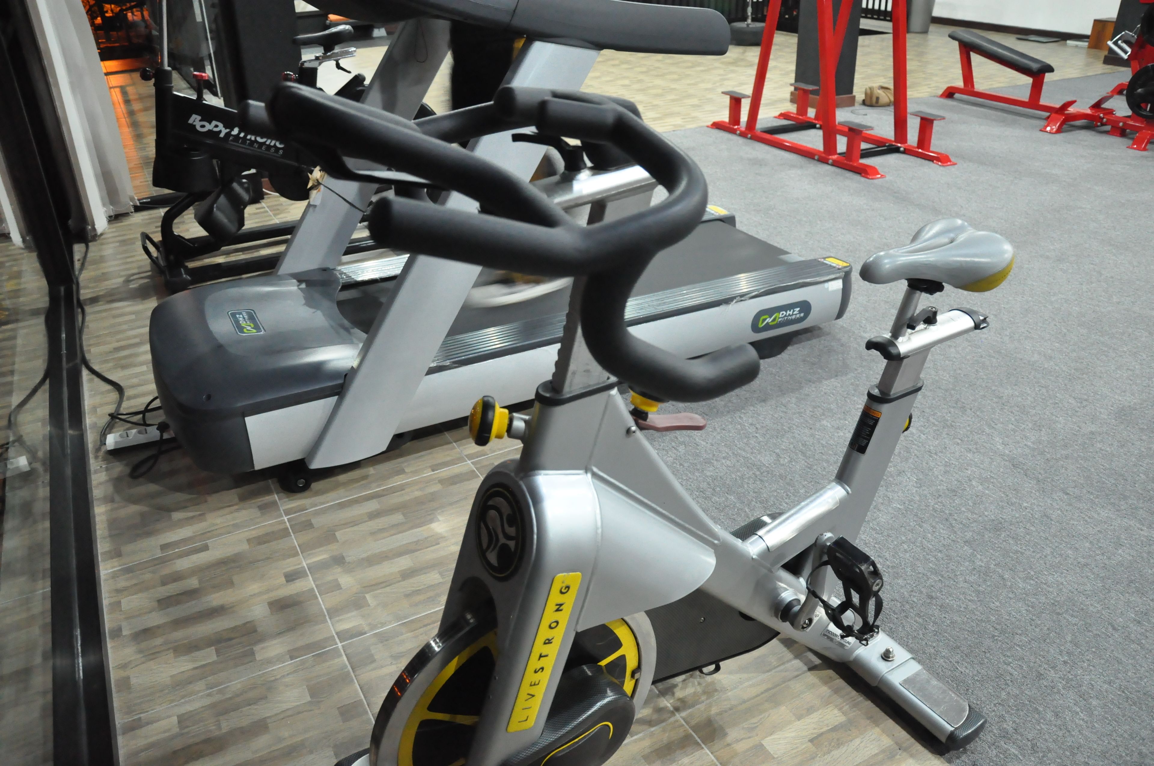 A stationary bike with a handlebar and a seat, located in a gym with a tiled floor and exercise equipment in the background.
