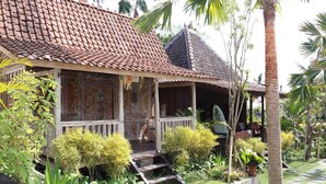 A traditional house with a tiled roof, a wooden porch, and a garden with various plants and a palm tree. - Kubudiuma Villas (Pererenan)