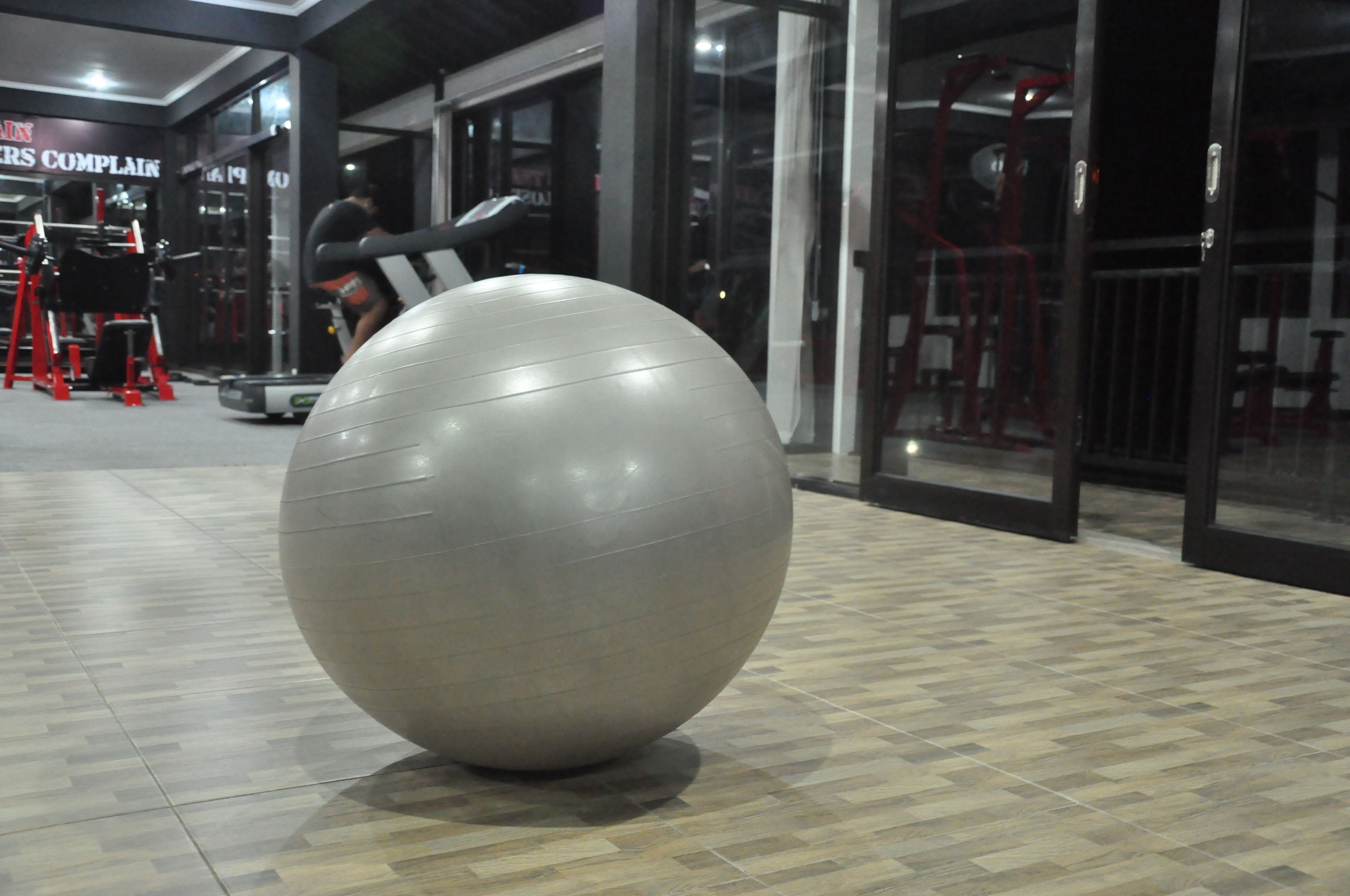 Ein großer grauer Trainingsball in einem Fitnessbereich mit Trainingsgeräten und einem Schild mit der Aufschrift 'PERS COMPLAINT'.