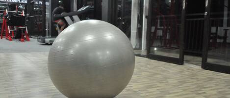 Ein großer grauer Trainingsball in einem Fitnessbereich mit Trainingsgeräten und einem Schild mit der Aufschrift 'PERS COMPLAINT'.