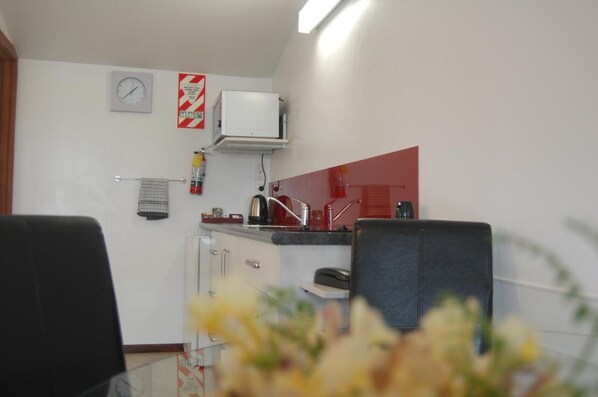 A small kitchen with a microwave, a kettle, and a sink. There is a clock on the wall and a fire extinguisher mounted above the counter.