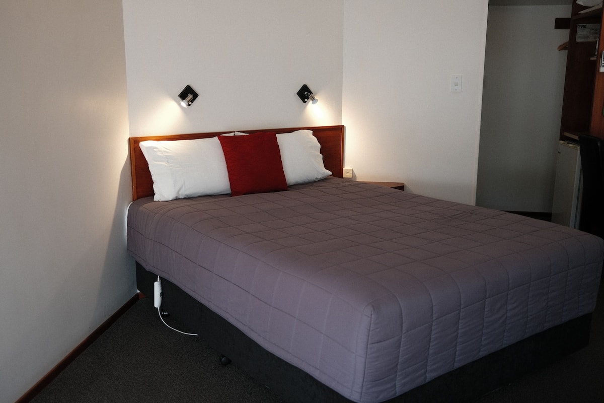 Un lit bien fait, avec une tête de lit en bois, un oreiller rouge et blanc, et deux appliques murales.