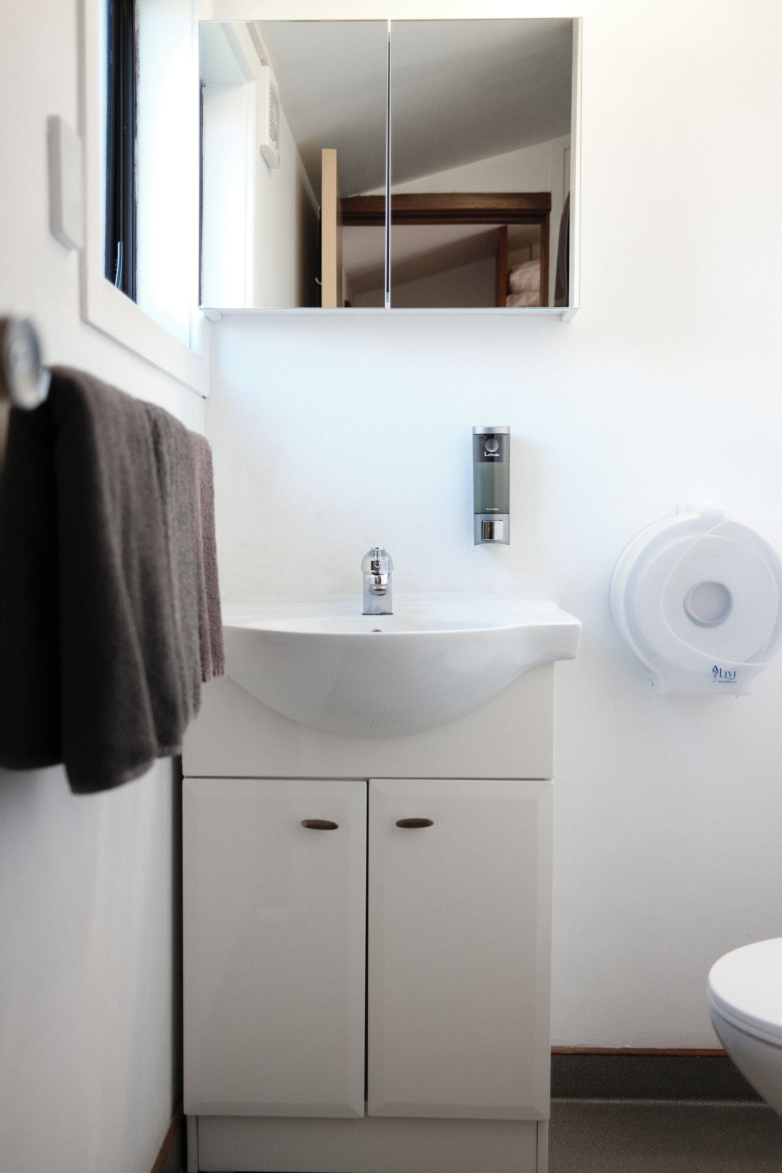Ein Badezimmer mit weißem Waschbecken, einem Spiegel, einem Handtuchhalter, einer Toilette und einem Papierhandtuchspender.