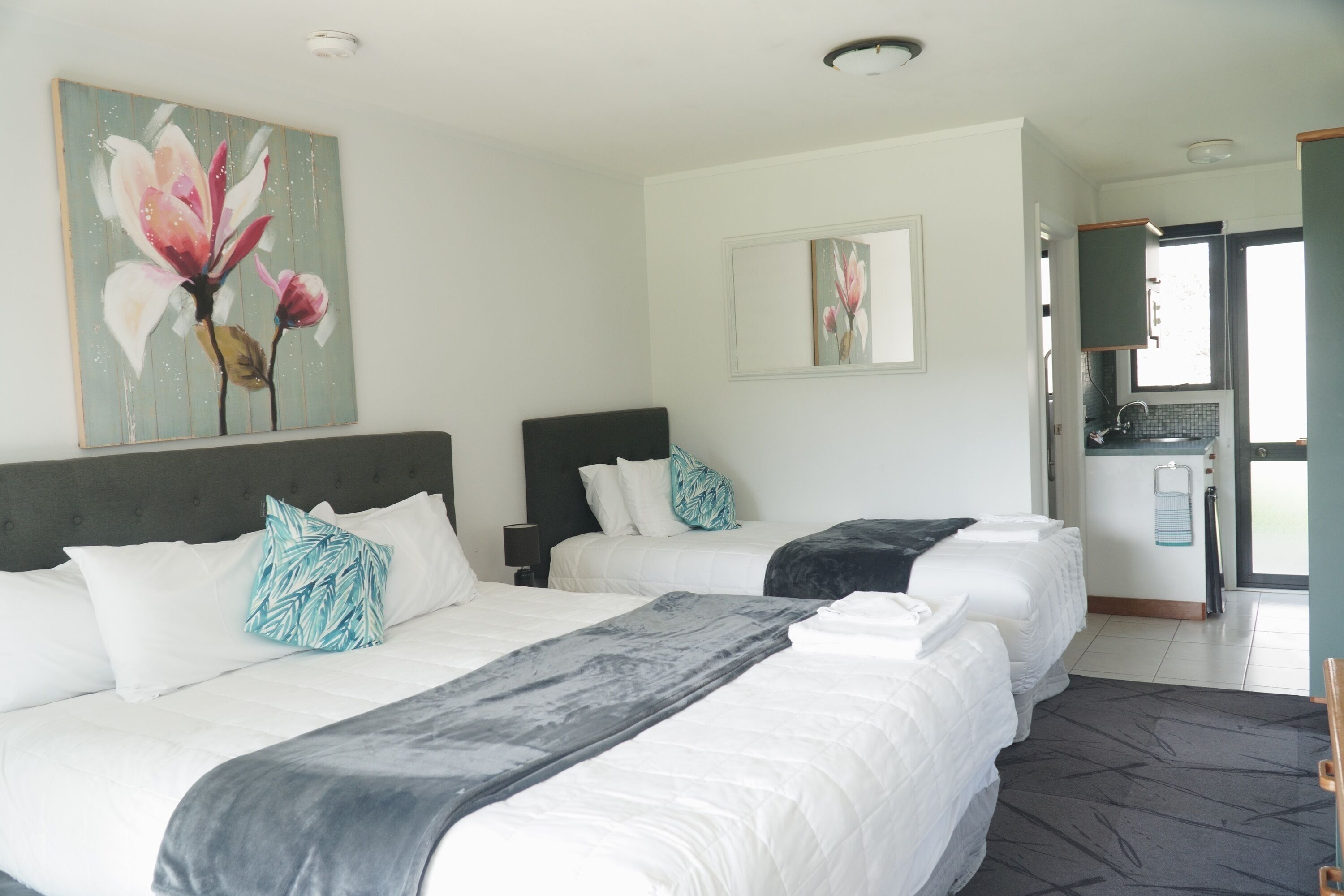 A hotel room with two beds, a painting of flowers, a small kitchen area, and a view of the outside.