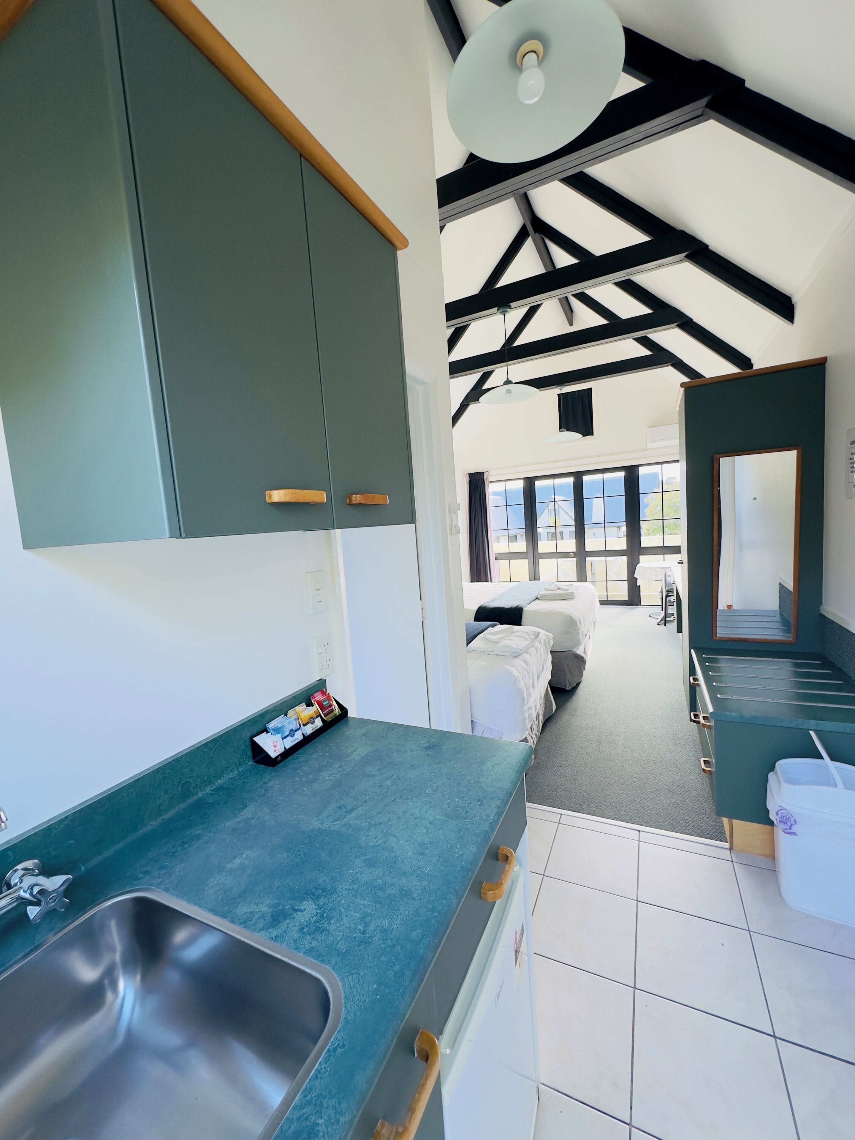 A modern kitchen with a stainless steel sink, green cabinets, and a view into a bedroom with a bed and a window.