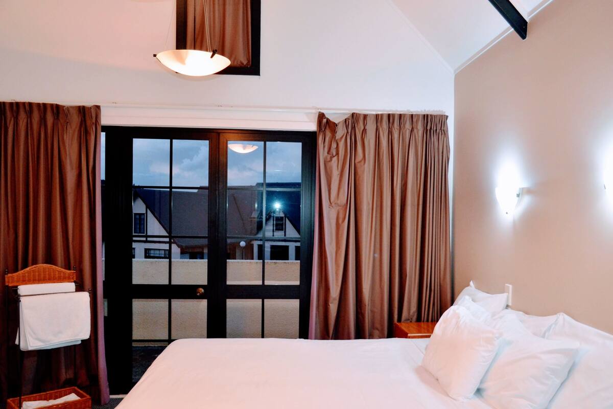 A hotel room with a large bed, a wooden headboard, a bedside table, and a view of a building through a glass door.