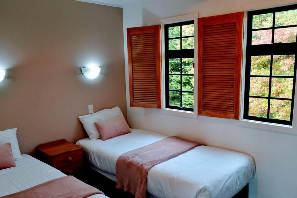 A room with two beds, a nightstand, and three windows with shutters.