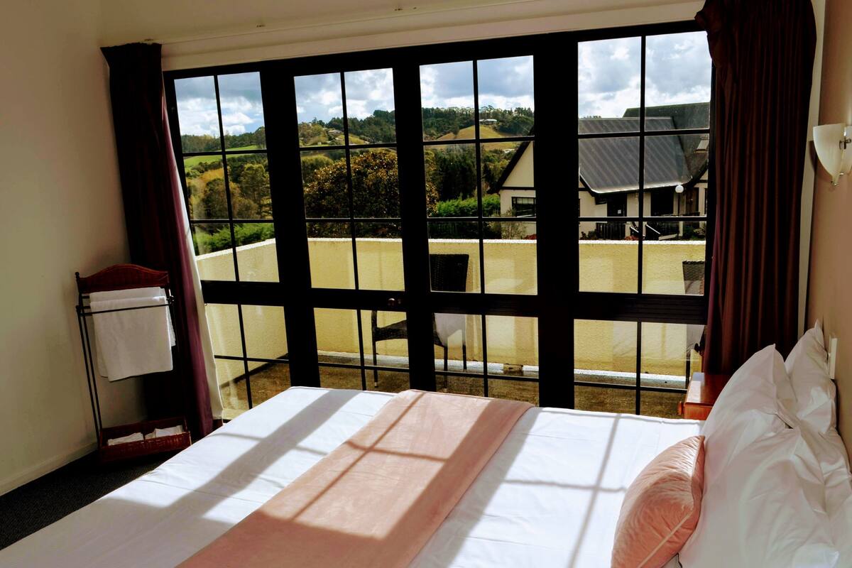 A hotel room with a large window, a bed with white linens, a wooden headboard, and a view of the countryside.