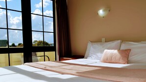 A hotel room with a large window, a bed with white linens, a pink throw, and a wall-mounted light fixture. - Salty Dog Inn (Snells Beach)