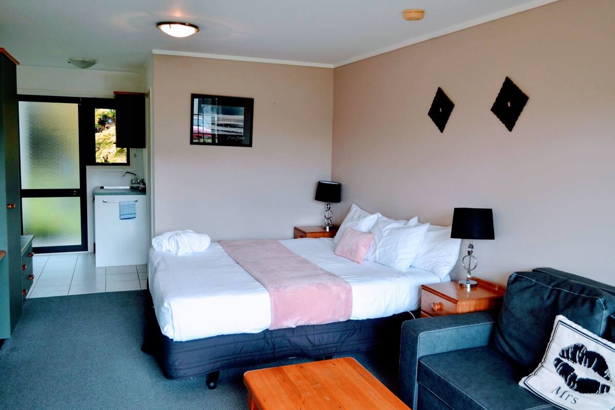 A hotel room with a bed, a sofa, a wooden table, a small desk, a lamp, a picture on the wall, and a window with a view.