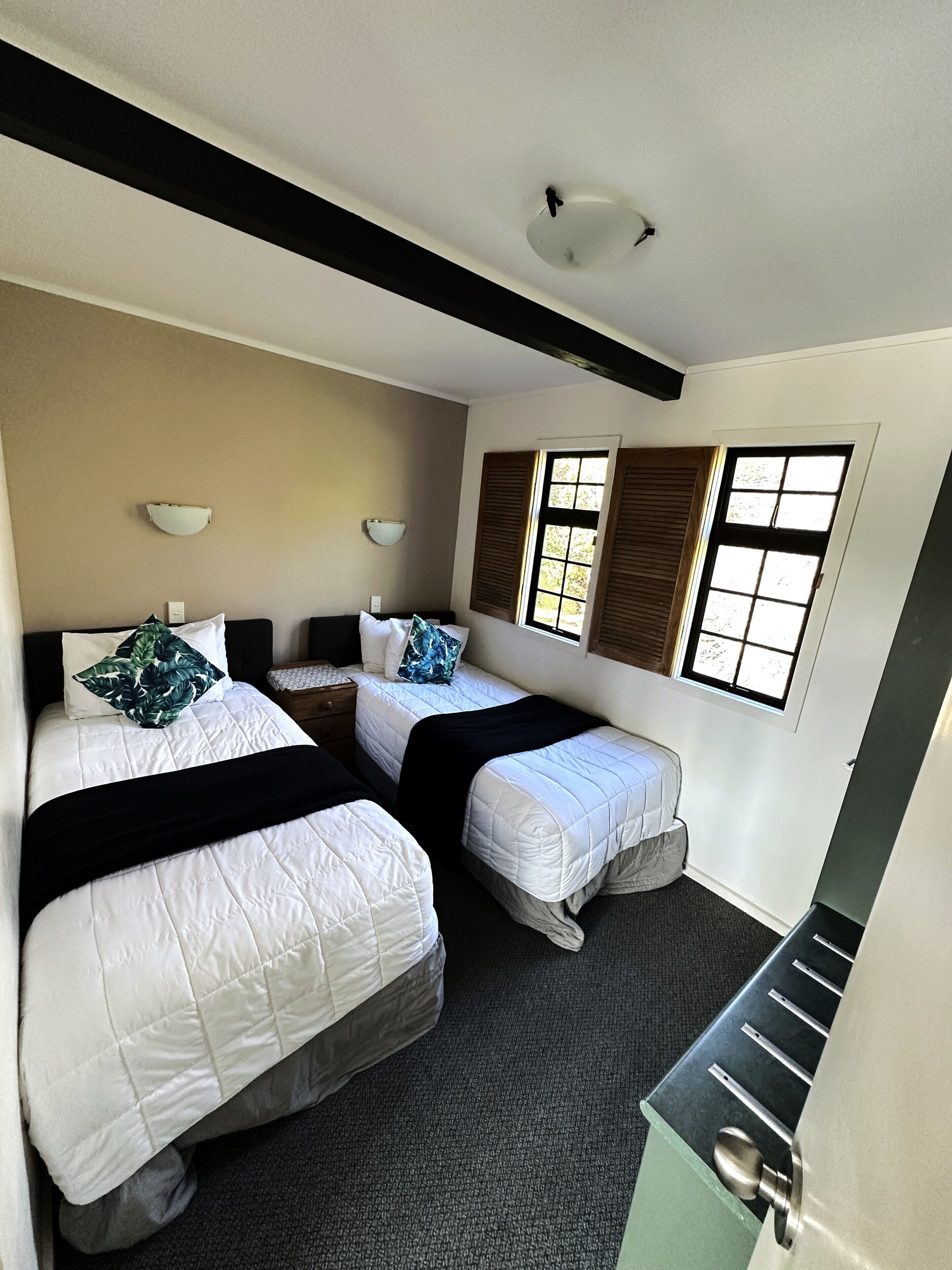 A hotel room with two beds, a window with shutters, a light fixture, and a small table.