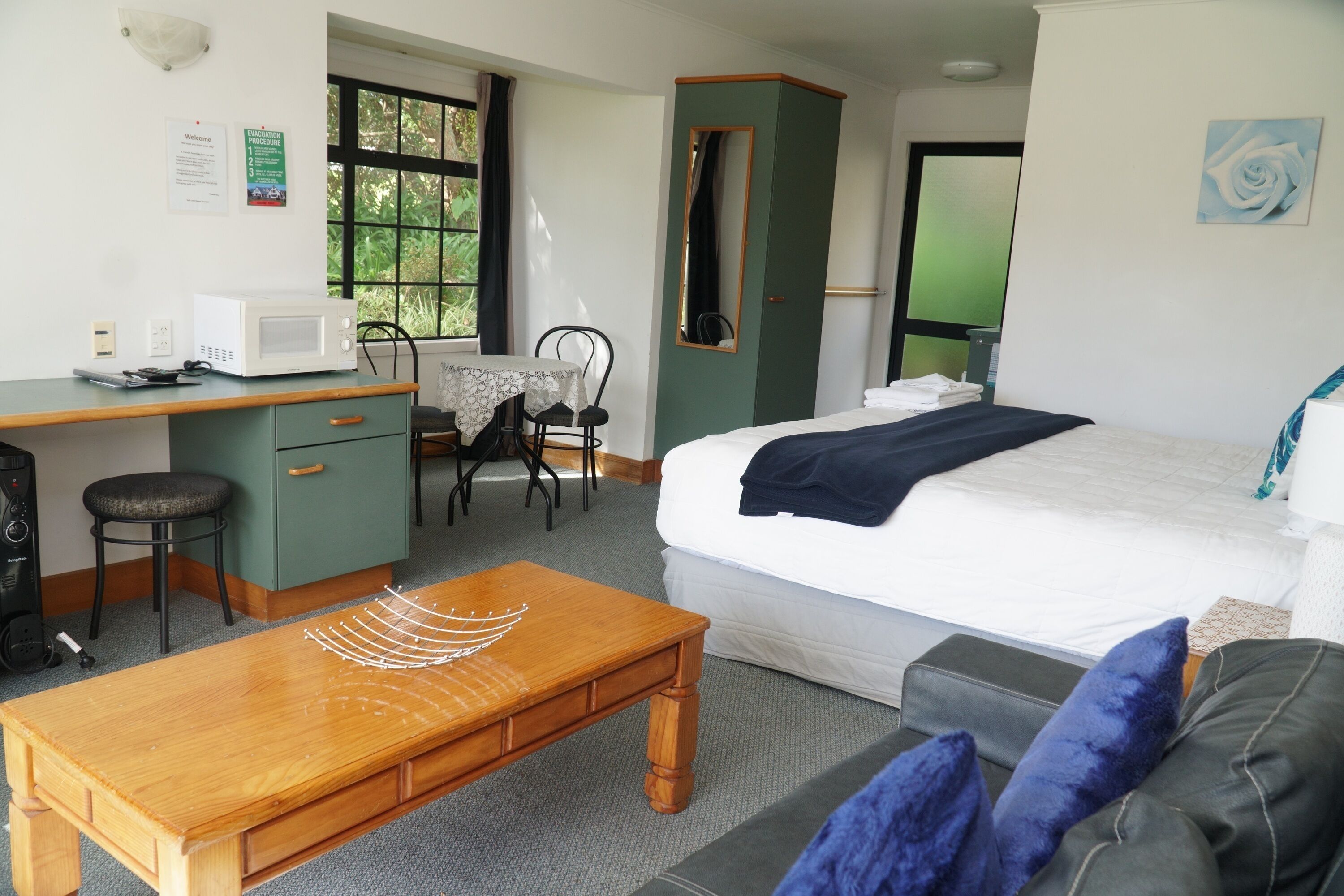 A hotel room with a bed, a desk with a microwave, a chair, a wooden table, and a window with a view of greenery.