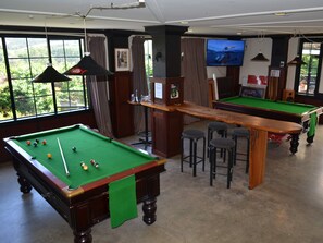 A pool hall with two pool tables, bar stools, a television, and large windows with a view of greenery. - Salty Dog Inn (Snells Beach)