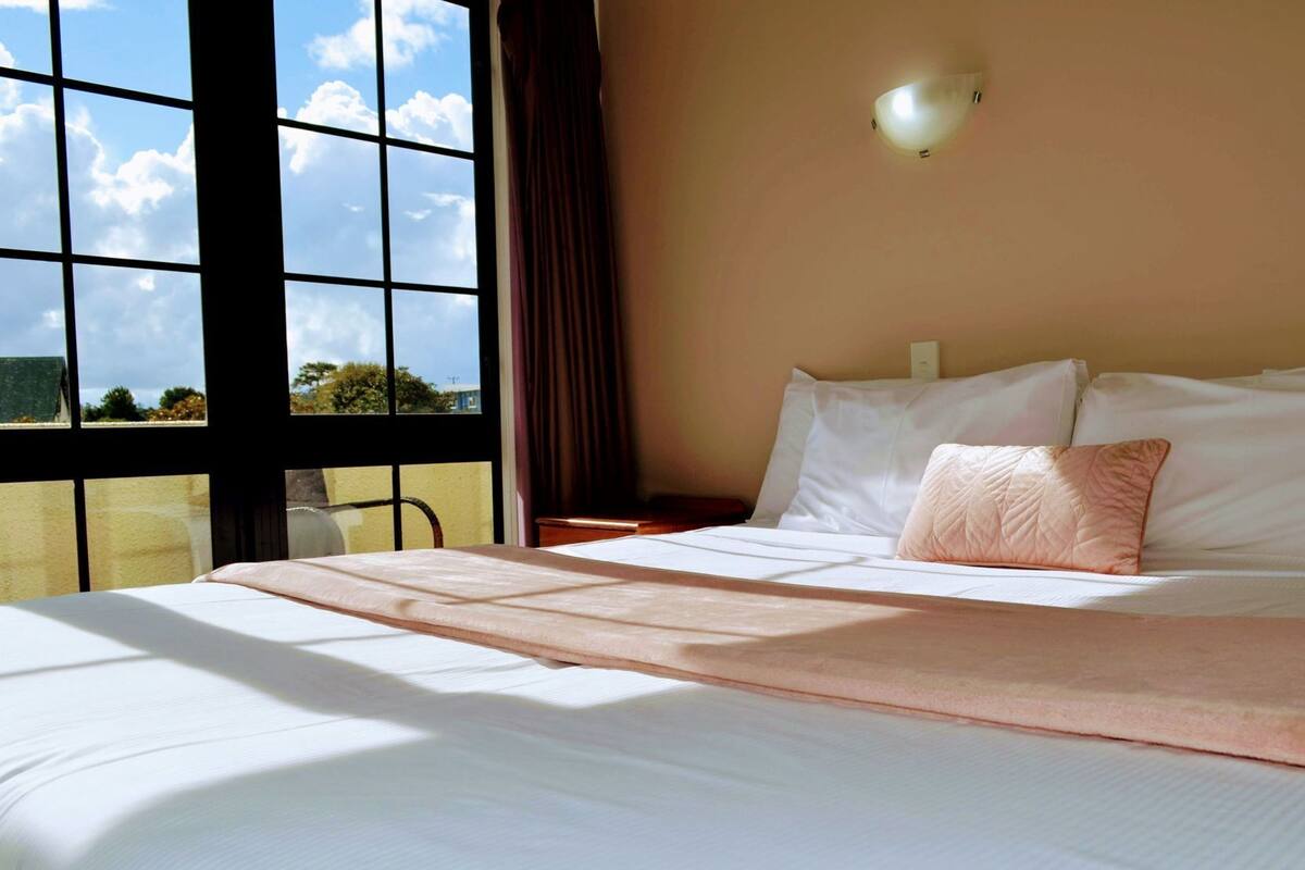 A hotel room with a large window, a bed with white linens, a pink pillow, and a wall-mounted light fixture.