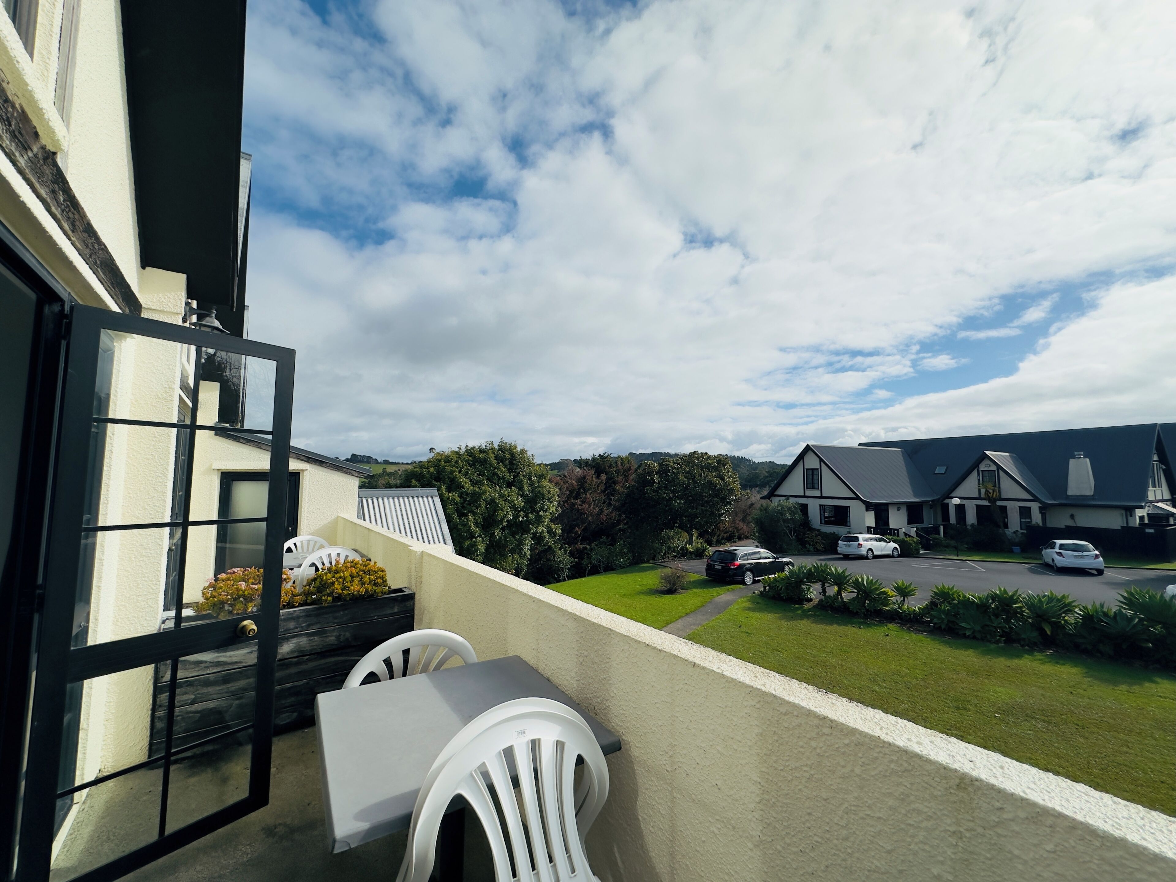 A balcony with a table and chairs, overlooking a parking area and houses.