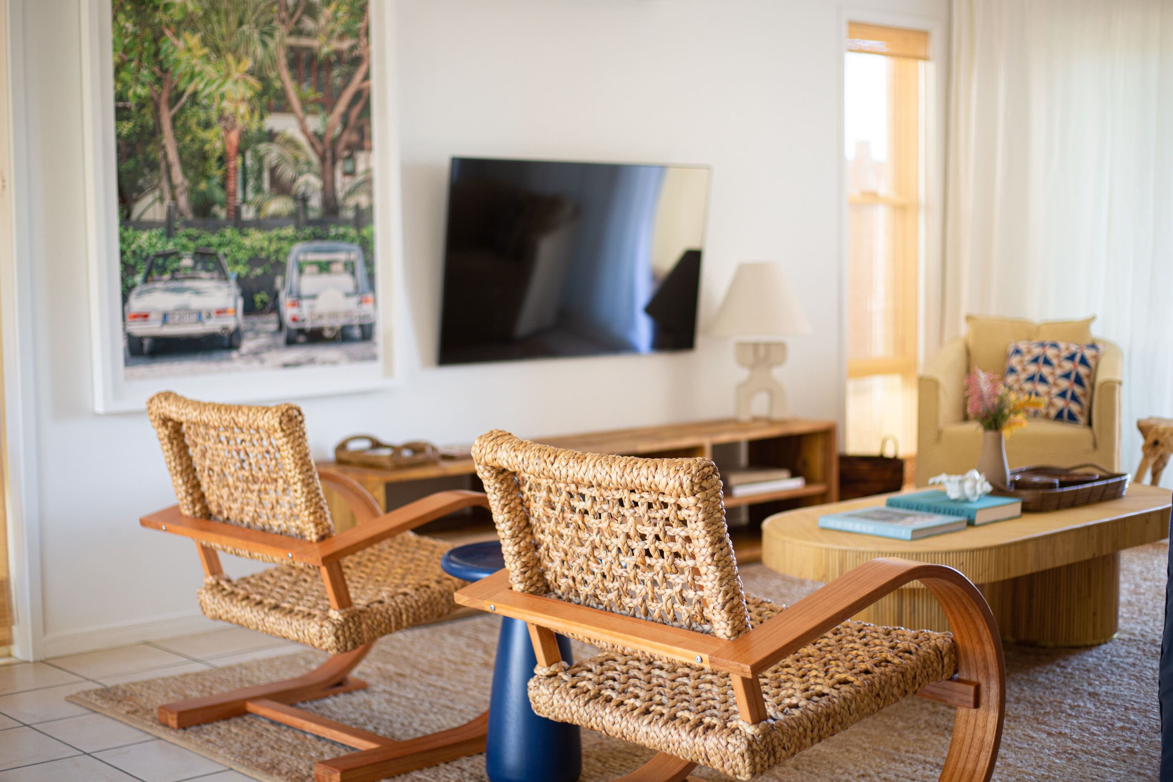 A living room with two wicker chairs, a coffee table, a television, and a window with a view of parked cars.