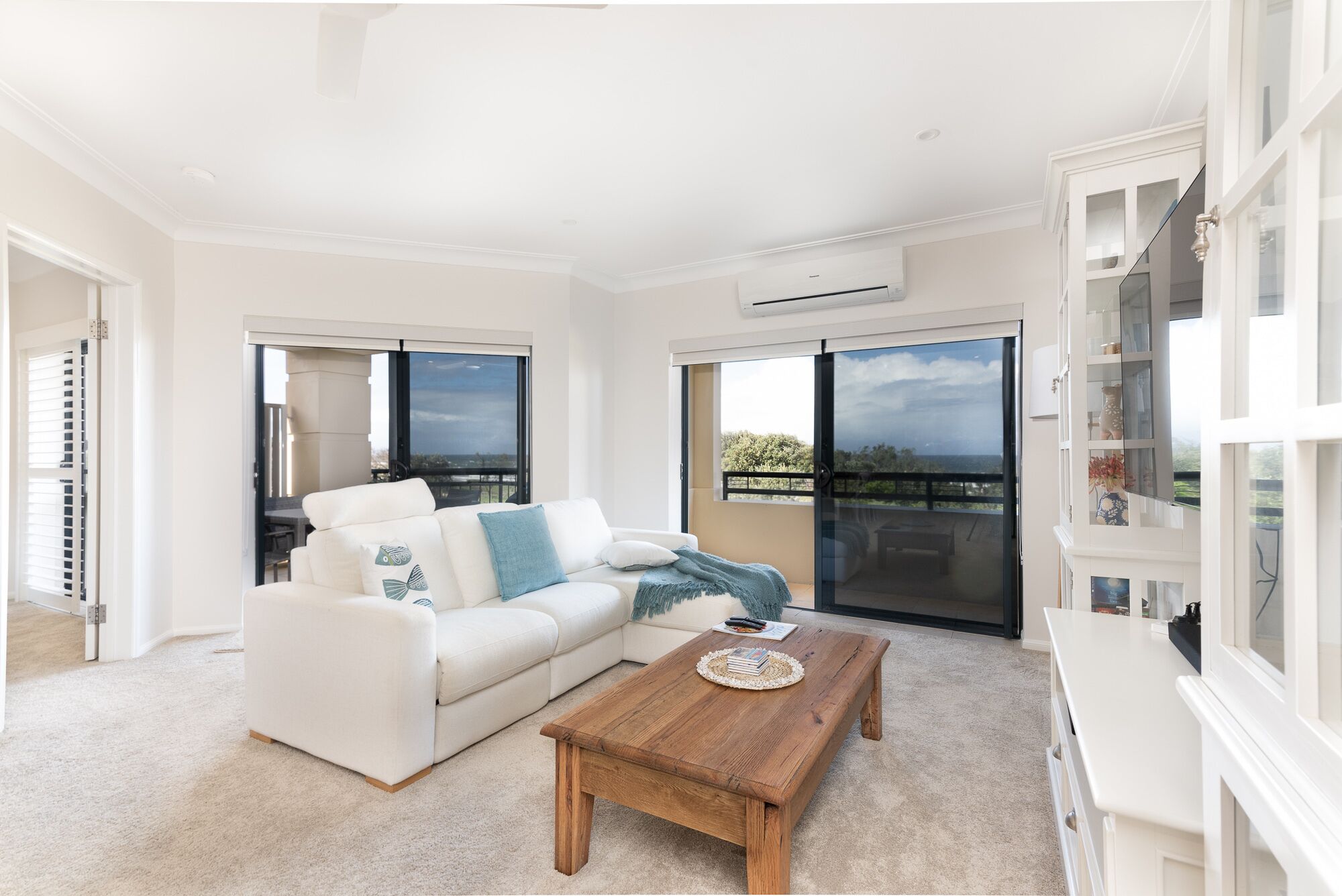 A modern living room with a white sofa, a wooden coffee table, and a view of the outdoors through large windows.
