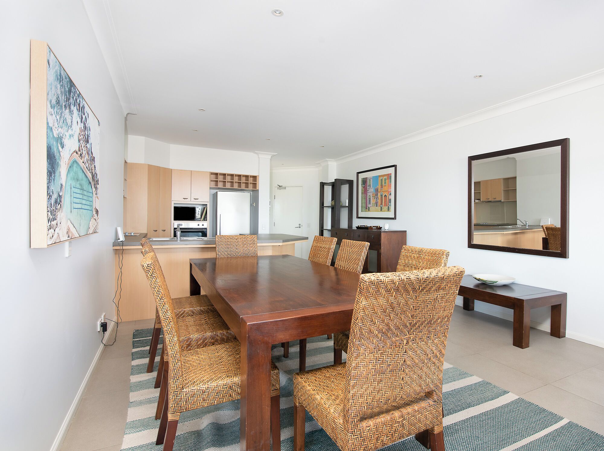 A modern dining area with a wooden table and woven chairs, a kitchen in the background, and a large mirror on the wall.