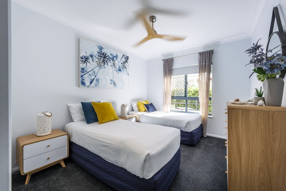 A bedroom with two beds, a ceiling fan, a window with curtains, a nightstand, and a dresser.