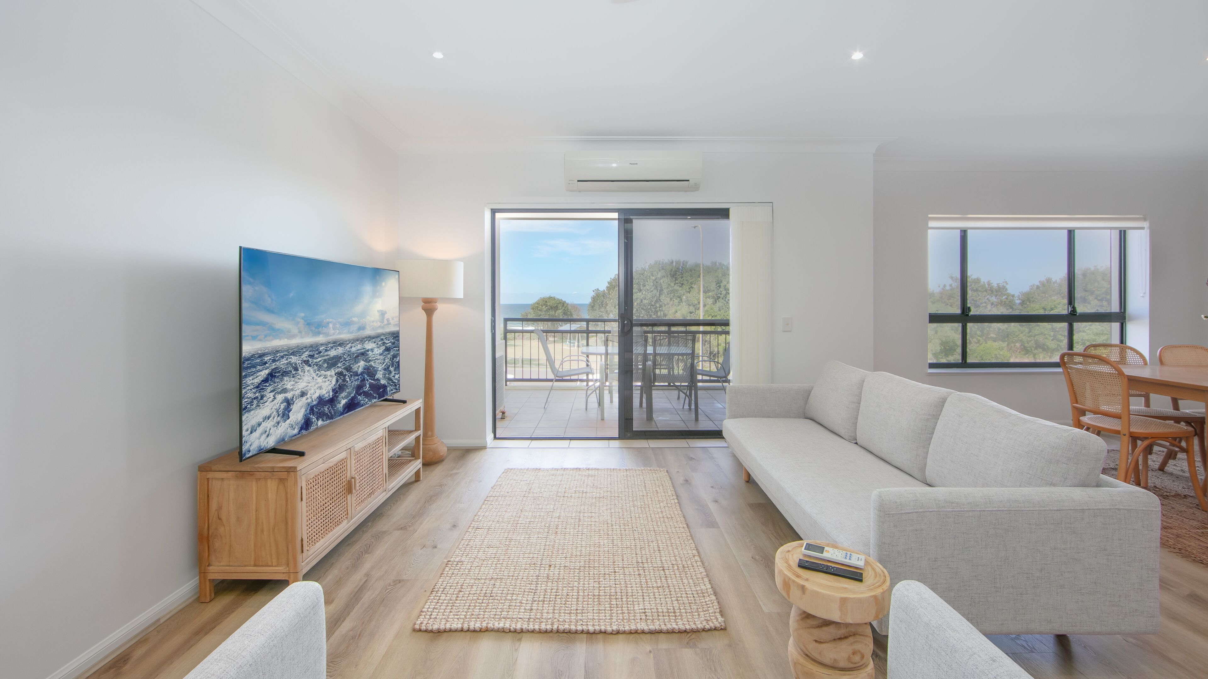A spacious living room with a large flat-screen TV, a sofa, a dining table with chairs, and a balcony with a view of the sea.