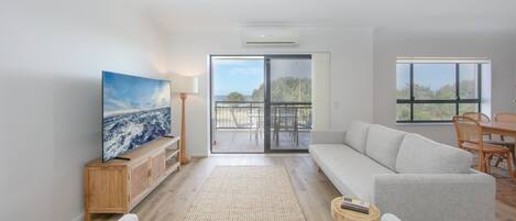 A spacious living room with a large flat-screen TV, a sofa, a dining table with chairs, and a balcony with a view of the sea.
