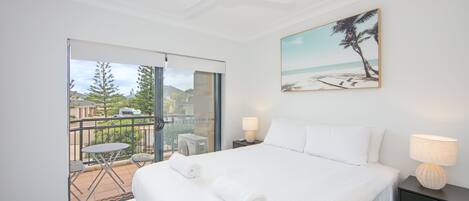 Une chambre d’hôtel avec un lit, deux lampes de chevet, un tableau représentant une plage et un balcon avec une table et des chaises.