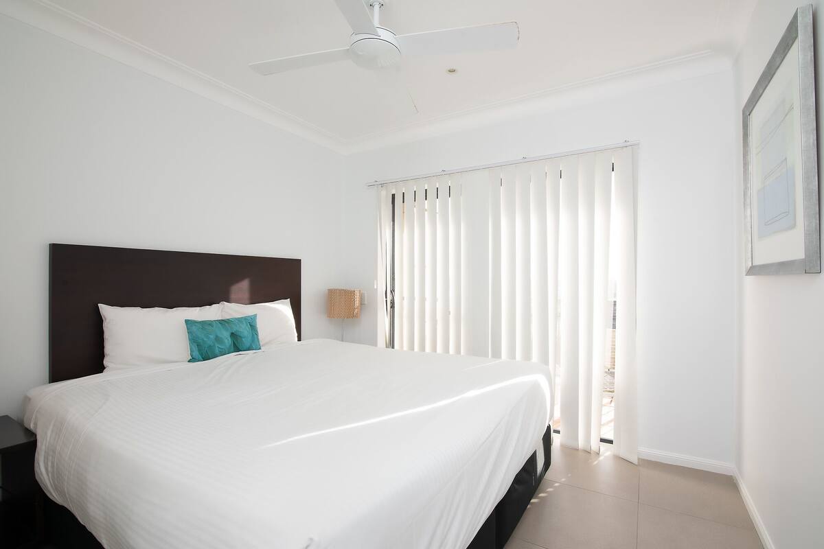 A bedroom with a large bed, a ceiling fan, and a window with vertical blinds.