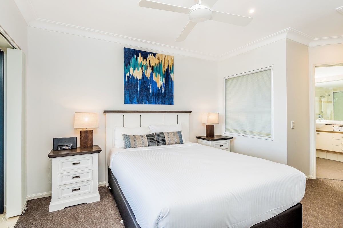 A bedroom with a bed, two bedside tables, a ceiling fan, a large window with blinds, and a wall-mounted abstract painting.