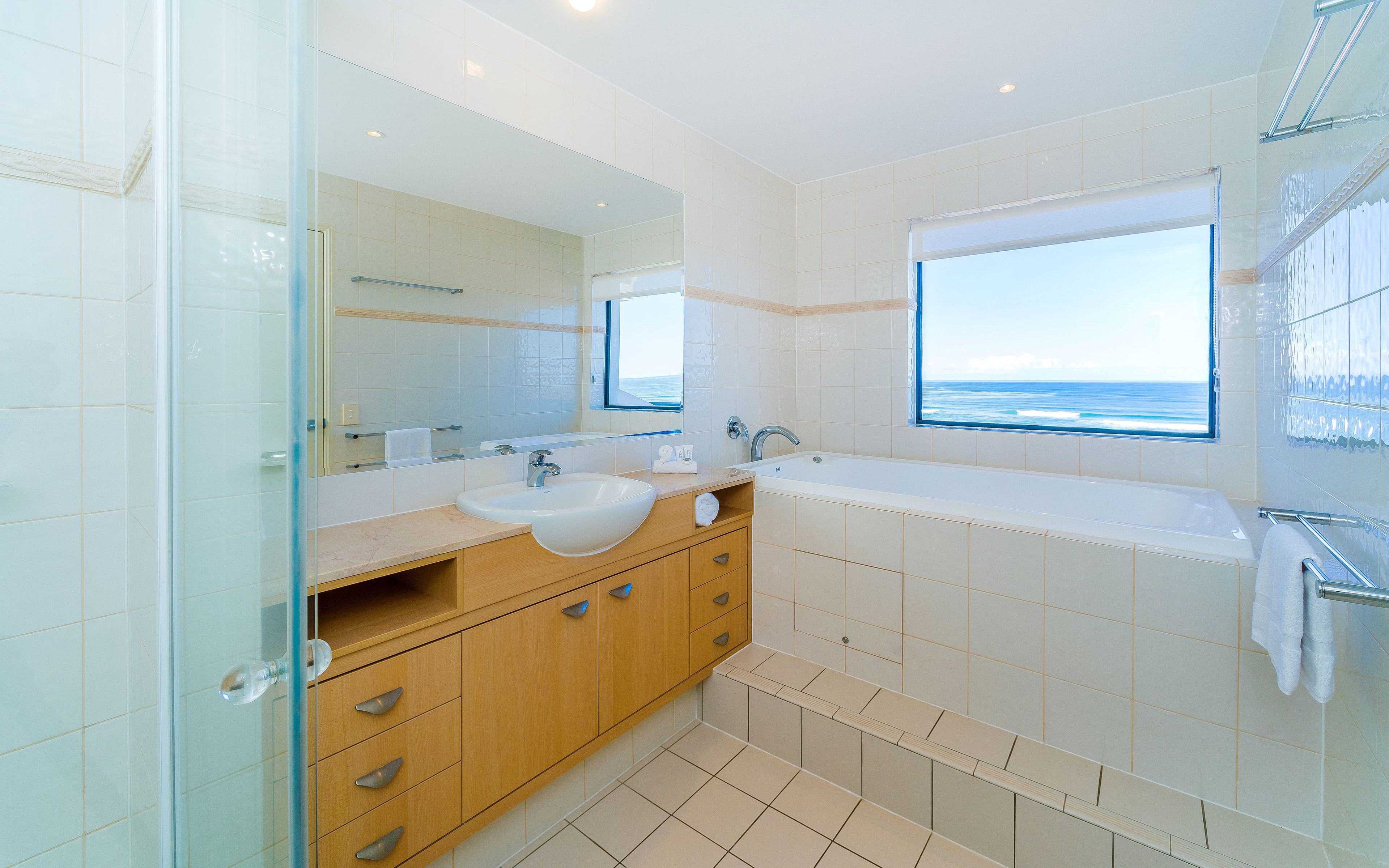 A modern bathroom with a large mirror, two sinks, a bathtub, and a window with a view of the ocean.