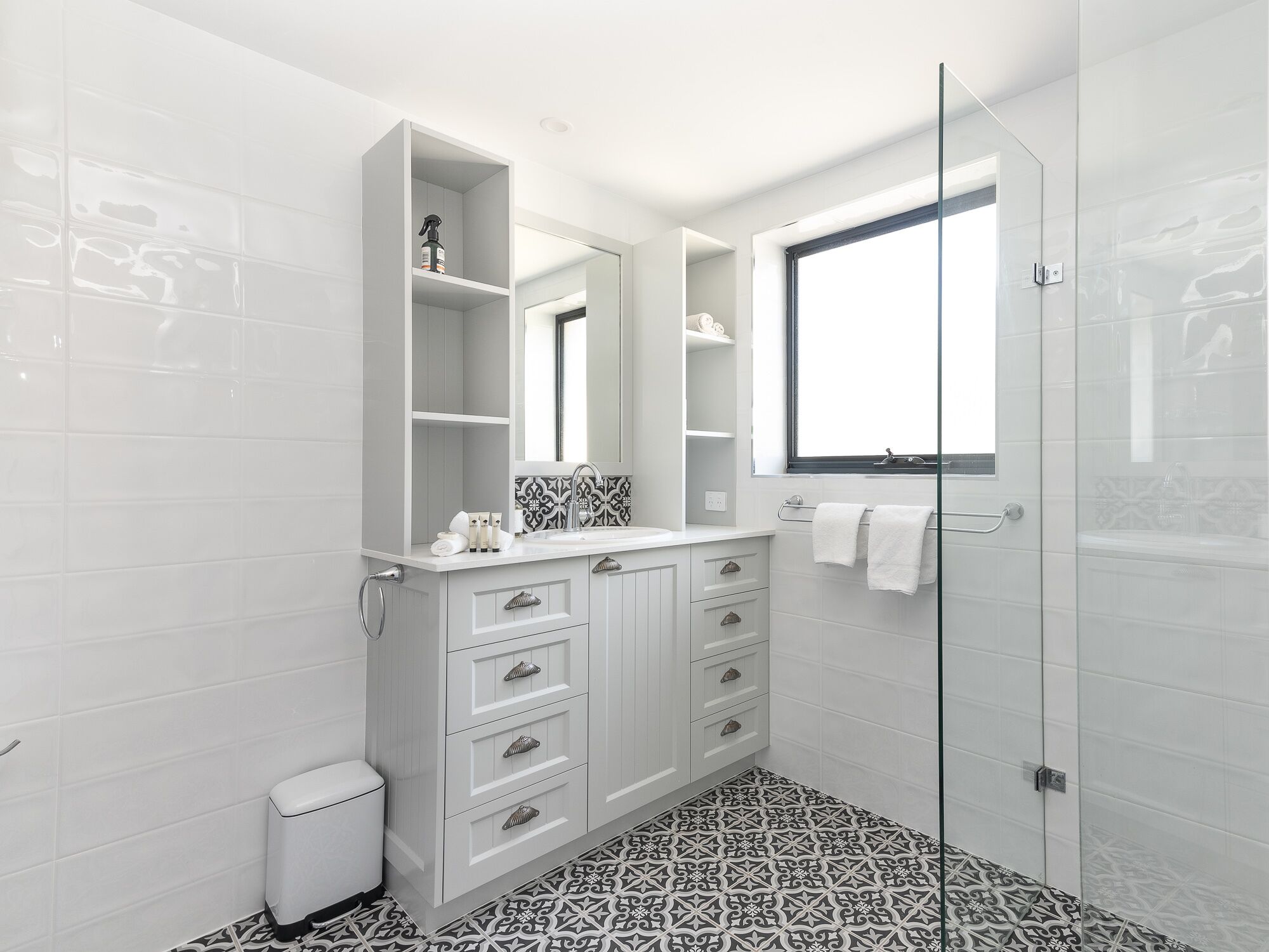 A modern bathroom with white cabinetry, a glass shower enclosure, and patterned floor tiles.