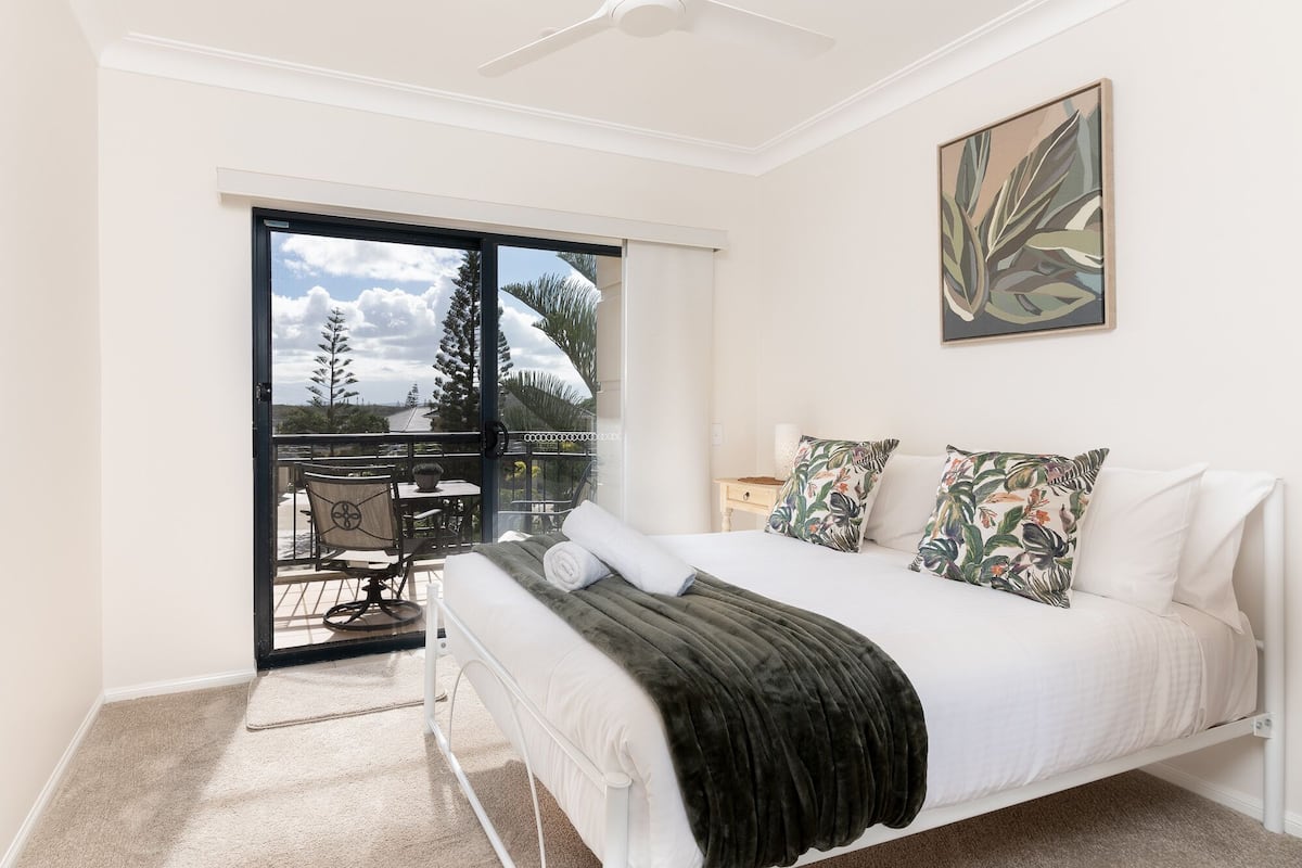 A bedroom with a bed, a ceiling fan, a balcony with a table and chairs, and a painting on the wall.