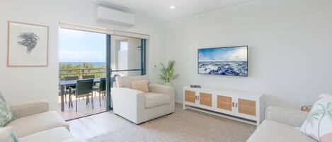 Een moderne woonkamer met een bank, een salontafel en een aan de muur bevestigde televisie. Er is een schuifdeur naar een buitenruimte met uitzicht op de zee.