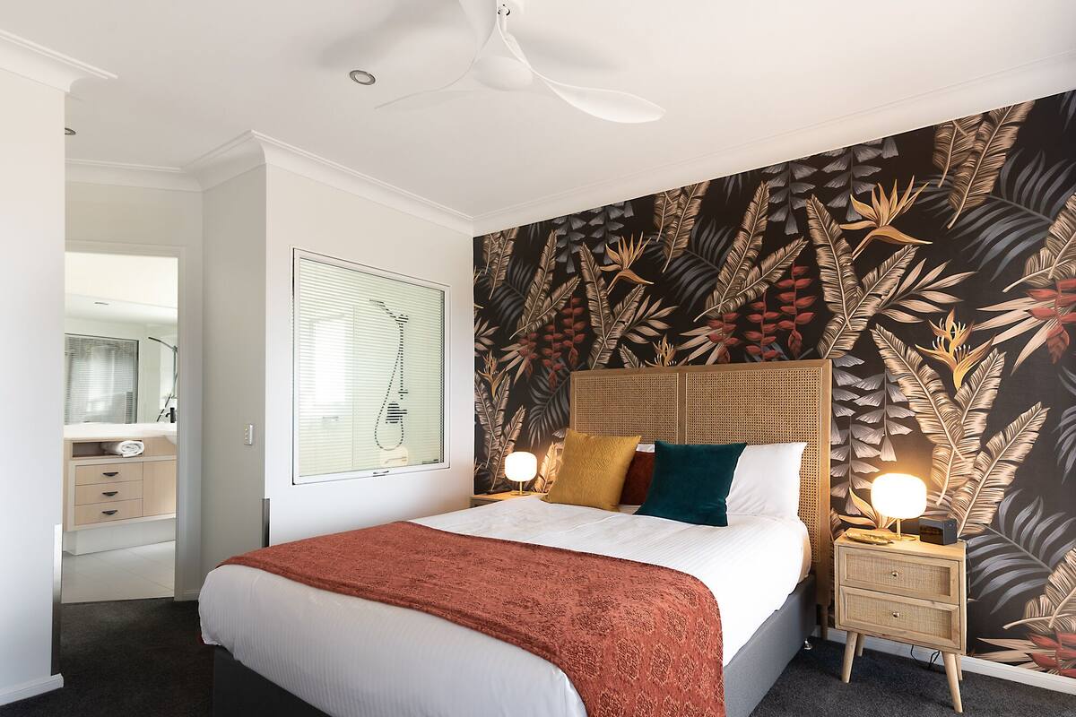 A bedroom with a bed, a nightstand, a mirror, and a wall with a tropical leaf pattern.