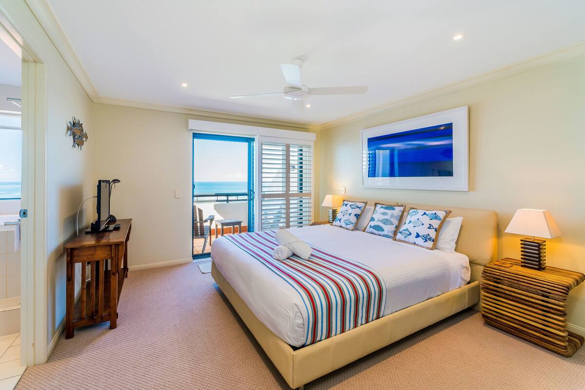 A bedroom with a bed, a desk, a chair, a lamp, and a view of the ocean.