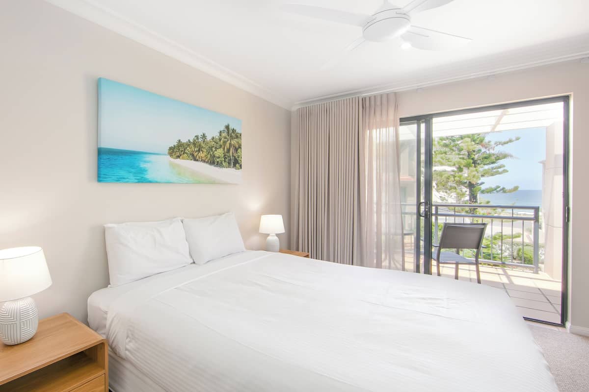 A hotel room with a bed, two bedside lamps, a painting of a beach, a balcony with a chair, and a ceiling fan.
