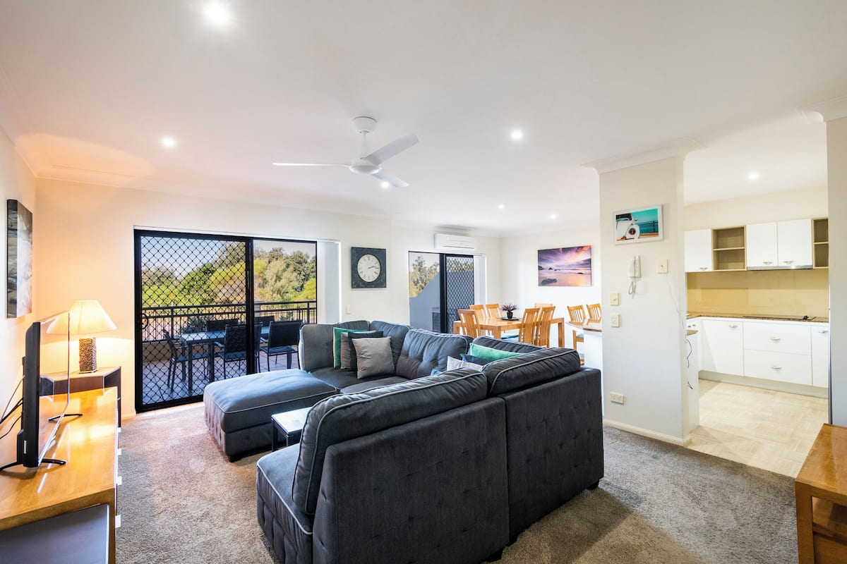 A modern living room with a sectional sofa, a dining area with a table and chairs, and a kitchen with white cabinets.