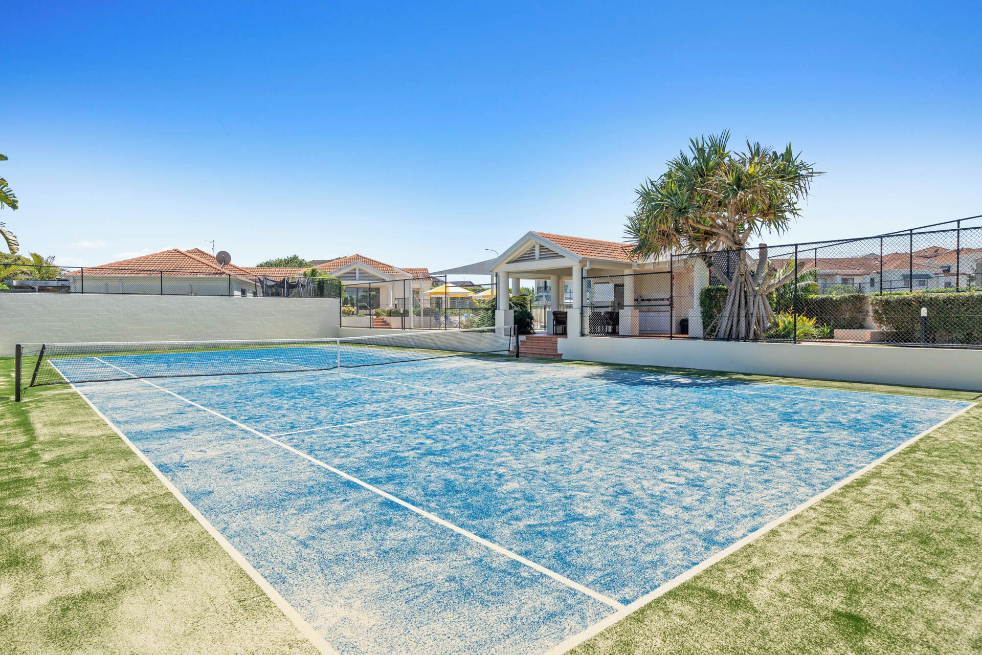 An outdoor tennis court with a net, surrounded by a fence and a building with a red roof.