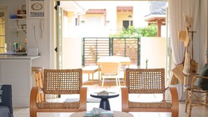 Una sala de estar con una mesa redonda de madera, dos sillas de mimbre, una mesa de comedor con sillas, un ventilador de techo y vistas al patio exterior.