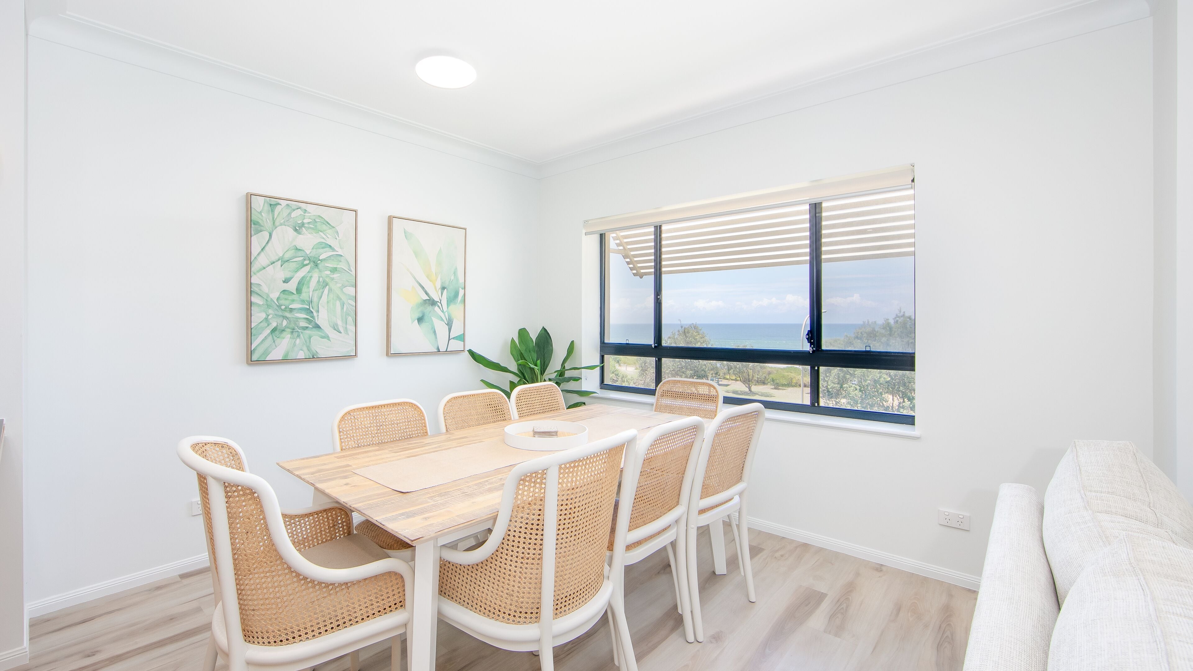 A dining area with a wooden table and wicker chairs, a potted plant, and a large window with a view of the ocean.